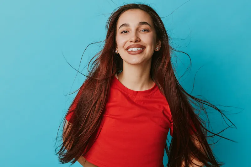 Young woman with long hair smiling and wearing red t shirt standing against blue background. Portrait of happy female with natural expression and casual style in studio lighting.