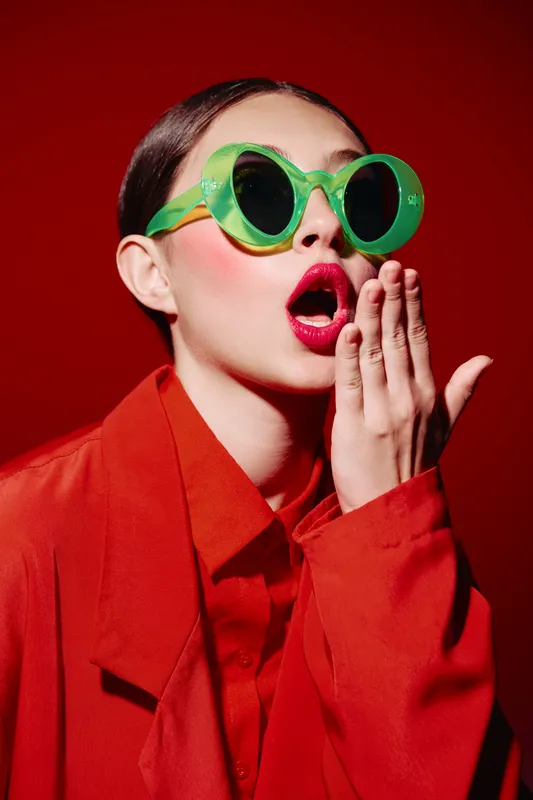 Young woman in bright red blazer wears round oversized green sunglasses. She covers her mouth with hand, expressing surprise in vivid makeup. Studio shot on red background, fashion and beauty concept.
