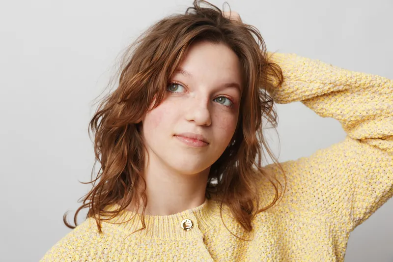 Young woman with wavy brown hair wearing a yellow sweater looks thoughtfully to the side. Studio portrait on a light grey background with natural expression and casual style.