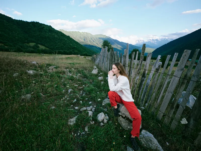 A woman leans on a rustic wooden fence in a wide alpine meadow, mountains in the distance, blue sky, creating a serene outdoor landscape perfect for nature lovers