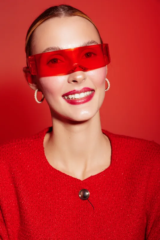 Smiling woman with red sunglasses and red sweater poses against red background. Fashion portrait with glossy lips and hoop earrings. Concept of style, confidence, and modern beauty.