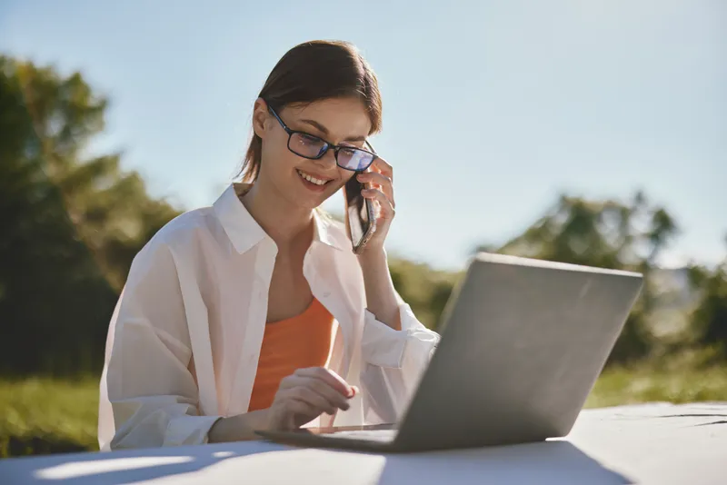 Cheerful young woman in glasses talking on the phone while working on a laptop outdoors, embodying productivity and a positive work life balance