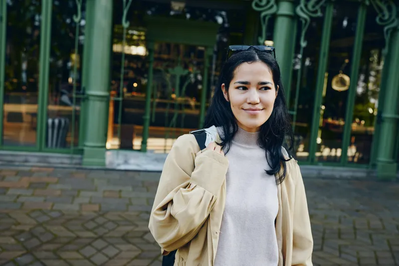 Woman outdoor fashion street casual backpack smiling jacket urban ambiance daylight candid scene on streetside with pedestrians and storefronts