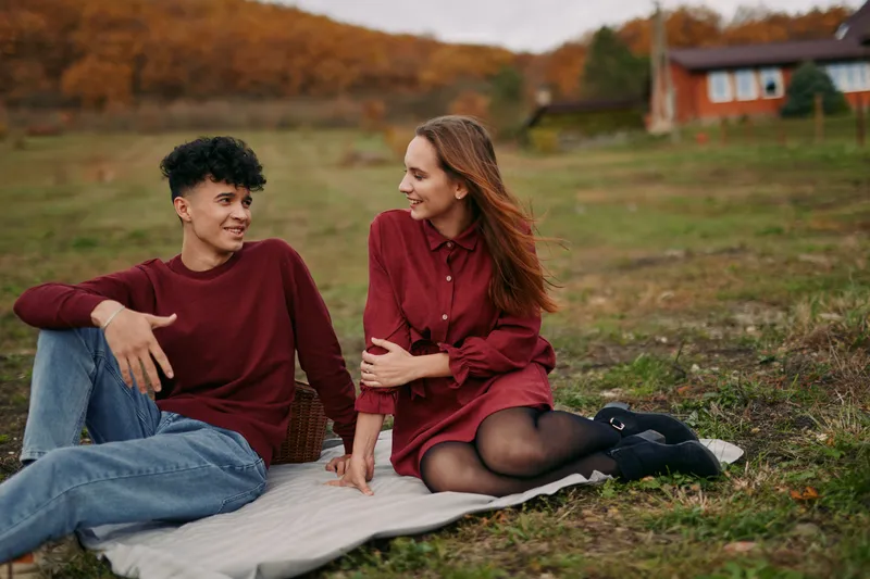A genuine candid portrait of a young couple sitting on a picnic blanket in a rural field, conveying authenticity and reliable connection through natural smiles and relaxed body language.