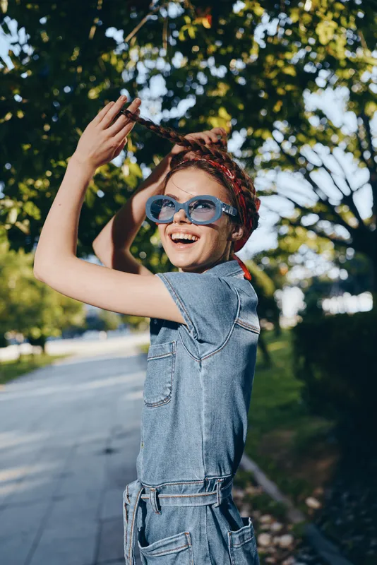 Lifestyle woman in modern boho-western denim street style wearing blue sunglasses joyfully plays with her braided hair outdoors in warm natural light film color effect.