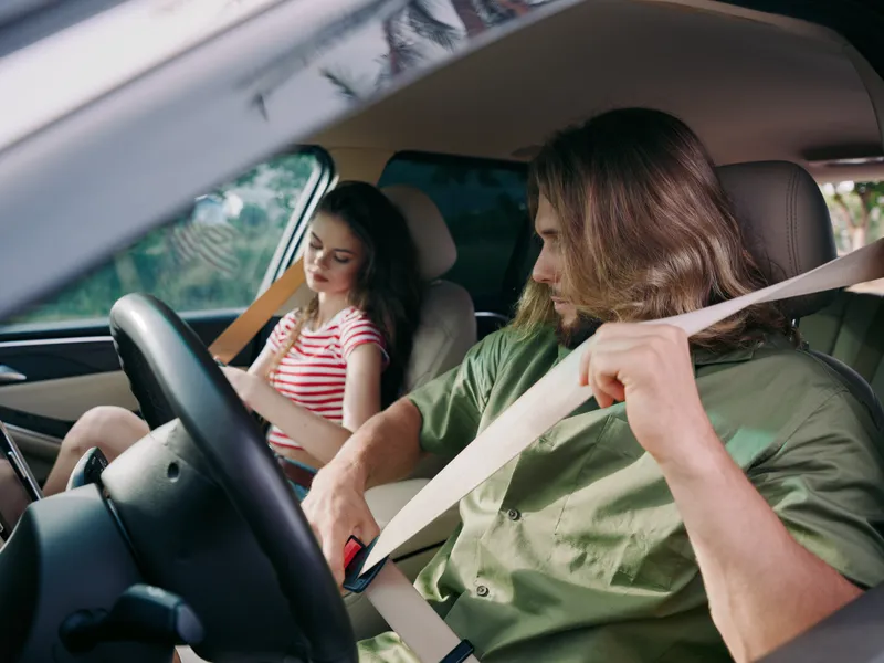 Two passengers in a car: a focused driver buckles his seatbelt while a calm young passenger sits in the back, enjoying a quiet road trip moment together.