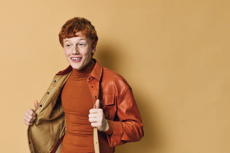 Redhead man with curly hair wearing brown leather jacket and turtleneck sweater smiling and looking aside on beige background. Person showing casual autumn fashion style.