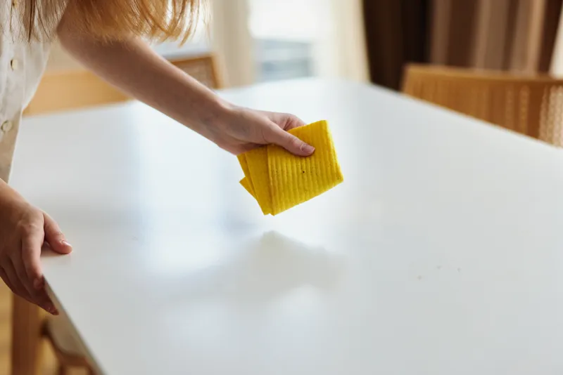 cleaning woman hand table sponge wipe kitchen home surface: Closeup of a female hand wiping a white dining table with a yellow sponge, domestic routine for tidy kitchen and household maintenance.