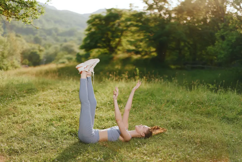 Young woman exercising outdoors in activewear, showing joy and relaxation with raised legs in a green, sunny landscape