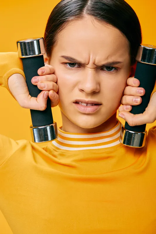 Fitness woman lifting dumbbells in a bold workout moment, showing strength and focus against a warm yellow backdrop. Intense expression communicates determination, effort, and sport performance