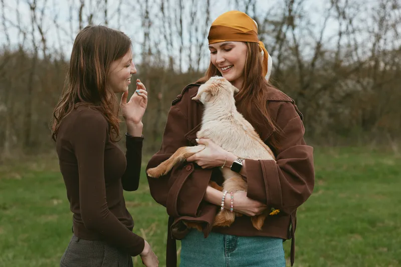 Outdoor scene with two women and a goat kid, warm interaction, joyful smiles, relaxed pose, natural colors, grassy meadow, soft breeze, lifestyle portrait, candid moment outdoors