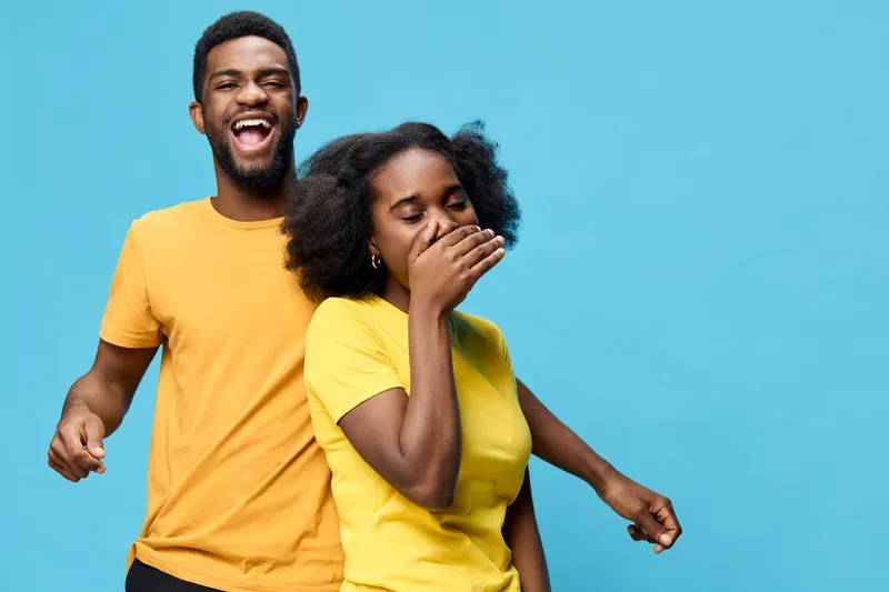A joyful young African couple laughing together against a bright blue background, wearing matching yellow shirts, embodying happiness and connection in a vibrant setting
