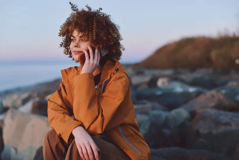 Woman lifestyle on a rocky seaside coast, wearing an orange jacket, curly hair lifted by wind, seated by ocean waves, soft daylight, calm thoughtful mood, outdoors nature travel portrait