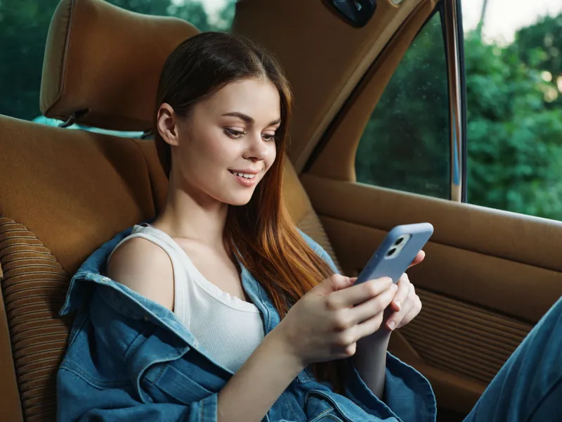 Young woman smiling while using a smartphone in a car, dressed casually in a denim jacket, capturing a moment of joy and connectivity in a natural setting.