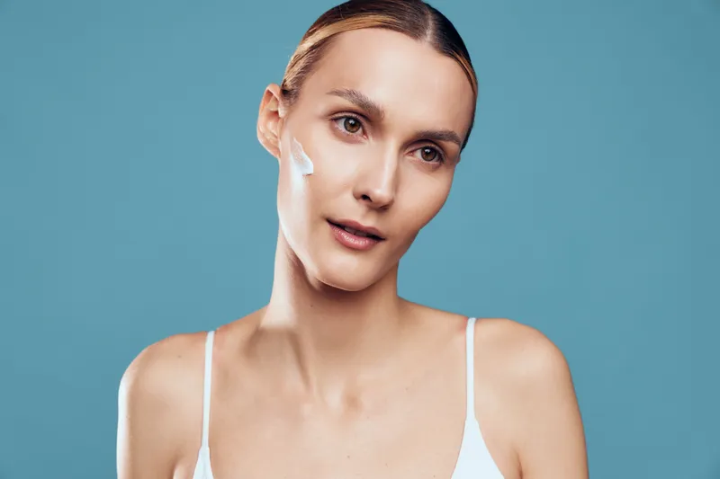 Young woman with clear skin applying white face cream on her cheek, wearing a white tank top against a blue background. Concept of skincare, beauty, and self-care routine.