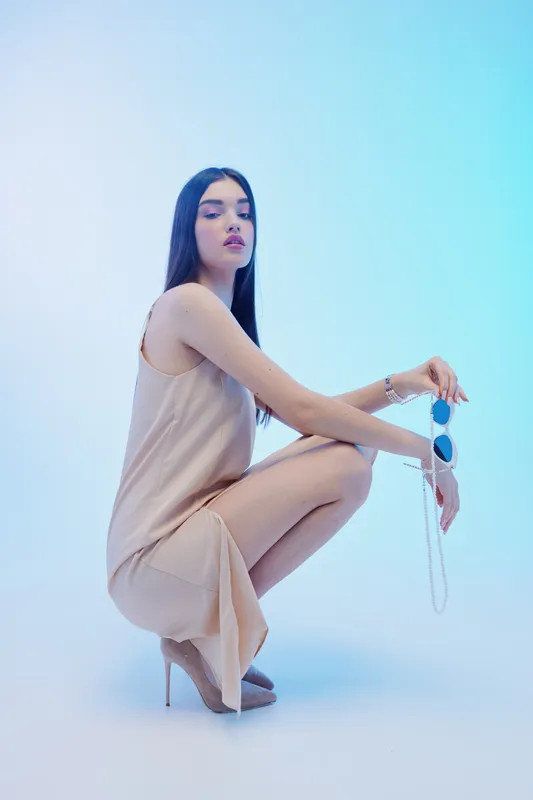Young woman in a beige dress and high heels poses holding sunglasses. Studio shot with soft blue background. Concept of beauty fashion and elegance.