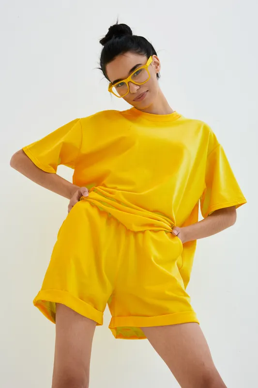Young woman wearing bright yellow casual outfit and matching glasses poses confidently against white background in stylish relaxed summer look with minimalist vibe.