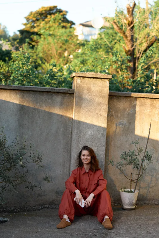 Cheerful woman outdoors, sitting on the ground in a red outfit, sunlit courtyard vibe, relaxed pose, warm tones, soft breeze, greenery background