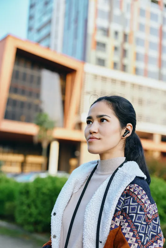 Woman outdoors in a city setting, stylish in a layered jacket and scarf, standing beside modern buildings, captured in natural daylight, showcasing urban fashion and confident mood