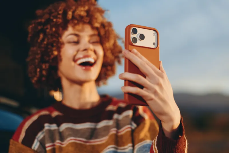 Woman selfie phone lifestyle outdoors laughing, curly hair close up portrait, she holds smartphone with orange case, golden evening light, travel feeling, soft bokeh background