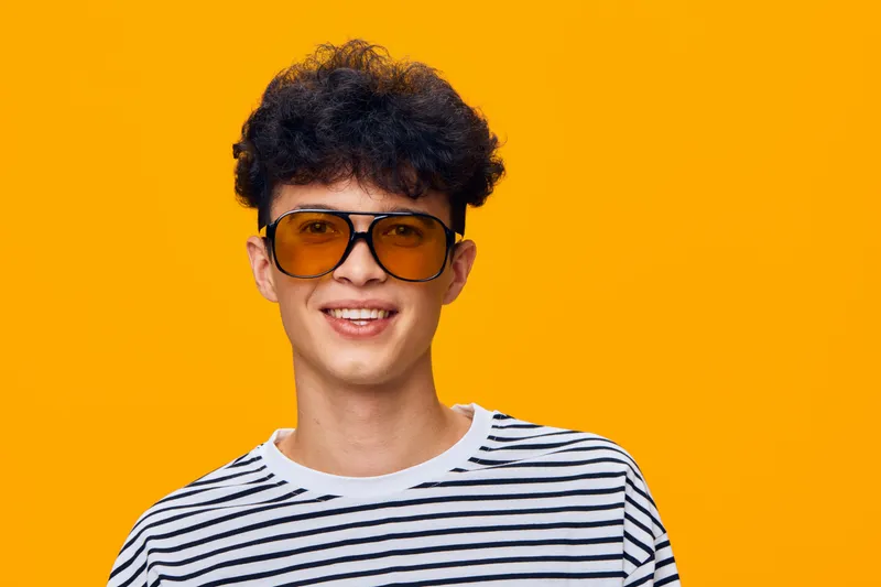 Smiling young man with curly hair wearing striped shirt and orange sunglasses on bright yellow background, showcasing joyful and trendy summer style.