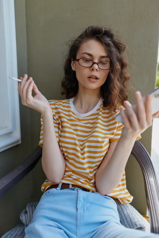 A young woman with glasses sits on a balcony wearing a yellow striped tee, smoking a cigarette and scrolling on her phone, capturing a casual urban mood