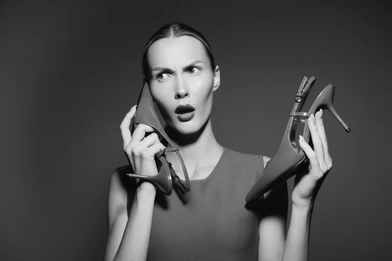 Confused Caucasian woman with pale skin holds pair of high heels near face in studio with grey background. Concept of indecision, fashion and shoes.