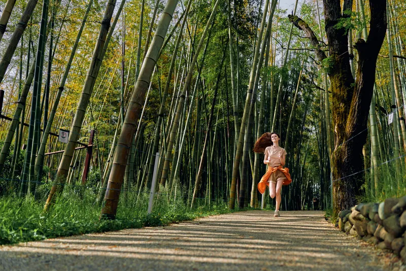 Woman running on gravel path through bamboo forest, orange dress flowing, motion and sunlight filtering through tall stalks, nature trail outdoor fitness in daytime joyful scene.