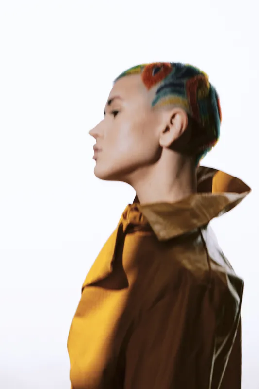 Woman with colorful short hair and floral design, wearing a mustard shirt, side profile with a serene expression on a bright white background, modern fashion portrait.