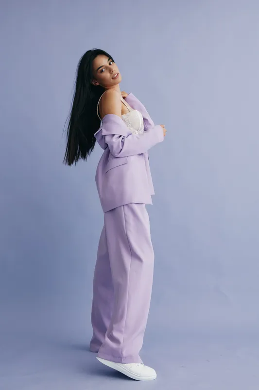 Young woman wearing lilac suit and white lace top stands confidently against pastel background, showcasing modern fashion and relaxed elegance in a stylish pose.