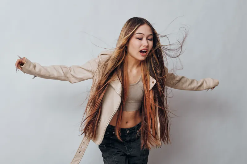 Young woman with long hair dancing and moving arms in beige jacket and black pants on gray background with expression and casual style.