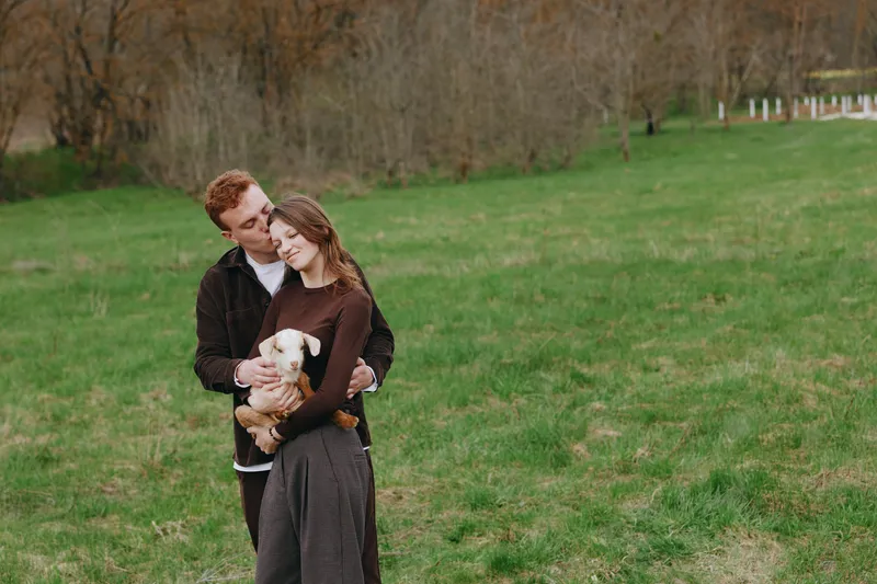 Young couple embraces tenderly in a green field as the woman holds a small white goat, sharing a quiet moment of affection and connection in a natural lifestyle setting.