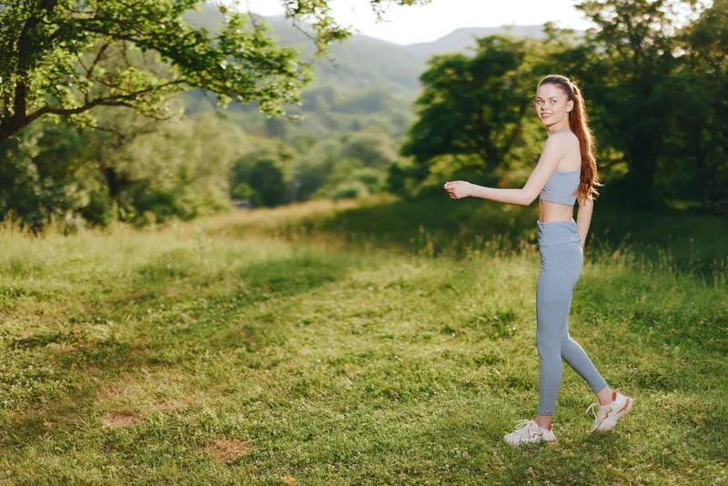 Happy young woman in activewear walking through a sunny green field, showcasing fitness and a healthy lifestyle with a joyful expression during a beautiful day outdoors