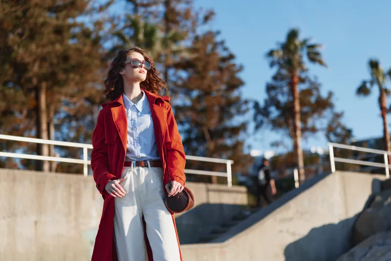 Stylish woman in a red coat over a light shirt and trousers stands outdoors, posing confidently under bright sun. Fashionable street style with bold color, relaxed elegance, and warm afternoon light.