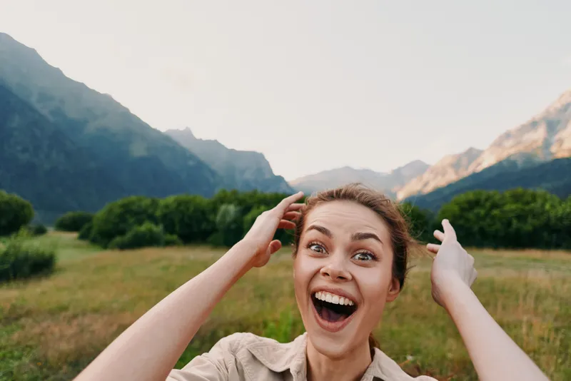 Young woman outdoors laughs with arms raised in a sunlit mountain valley, a candid moment of joy and freedom amid lush scenery