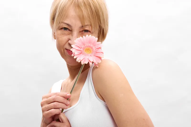 Blond woman portrait smiling closeup holding pink flower near face, soft light, white studio background, fresh spring mood, gentle expression, calm