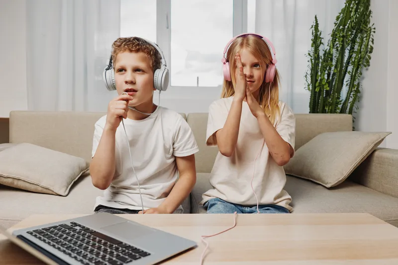 Two children wearing headphones sing and listen while sitting on a couch near a laptop in a bright living room.