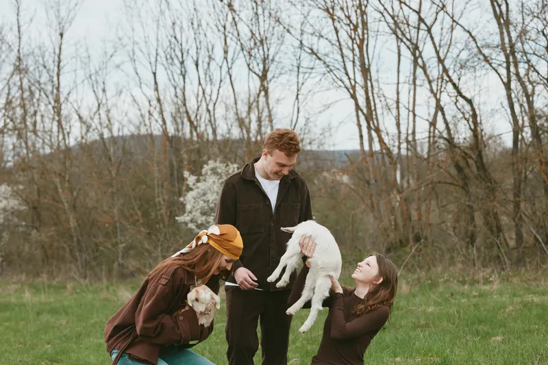 A young man and two young women enjoy a spring day outdoors while holding and playing with a white baby goat in a grassy field with bare trees in the background.