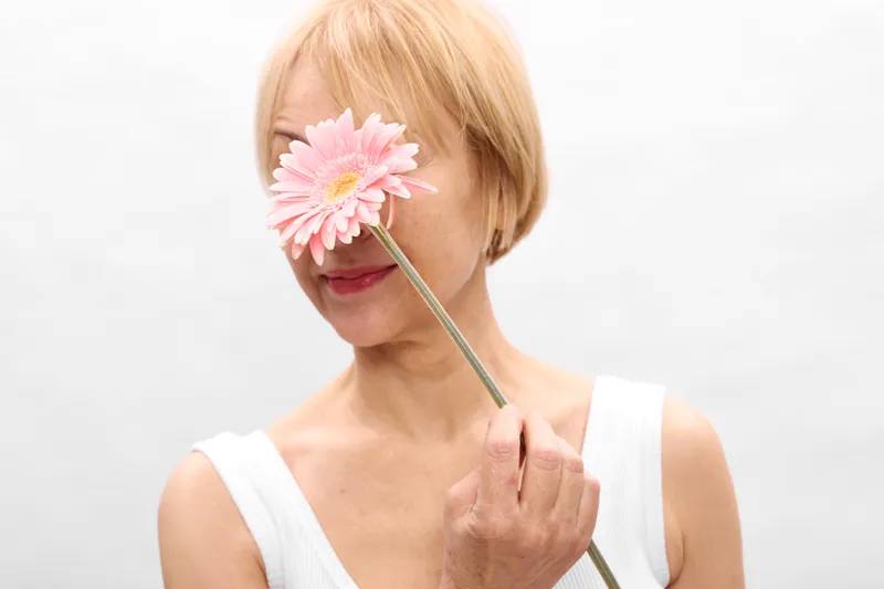 Woman portrait with blonde hair holding pink flower petals over her eyes, covering them with one hand, soft beauty in a white studio background for calm romance