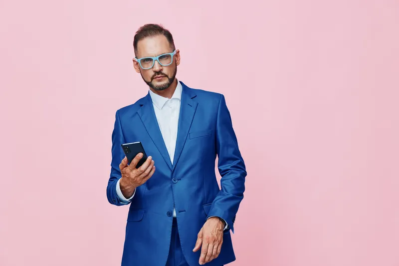 Professional man in a blue suit stands against a solid pink background, exuding confidence and focus while checking a smartphone. The isolated color backdrop highlights his poised, businesslike