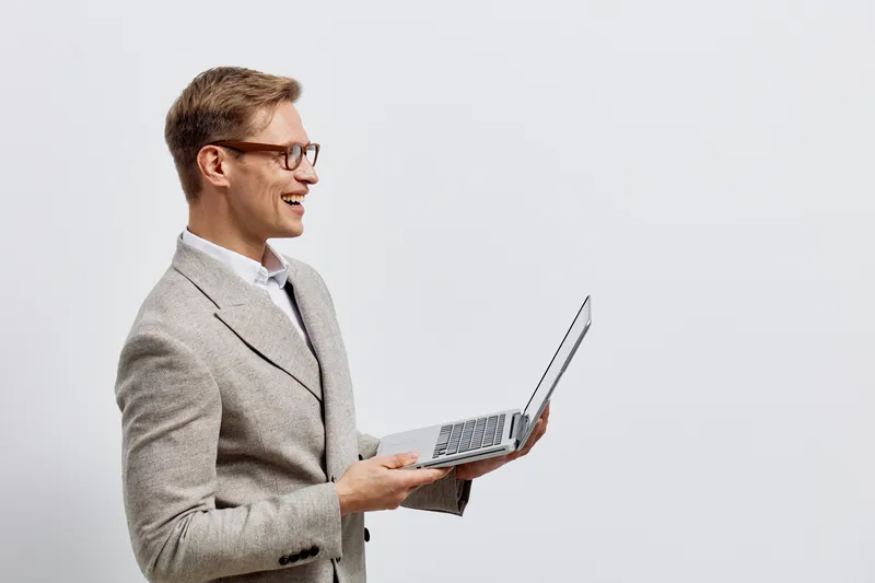 Smiling man in business casual holding a laptop computer, wearing glasses and blazer, isolated against plain white background. Office work, technology, professional, modern, confident, cheerful mood, 