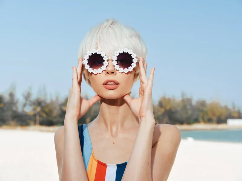 A stylish model with platinum short hair wears round flower-framed sunglasses at a sunny beach, creating bold summer fashion and a playful contrast against the blue sky.
