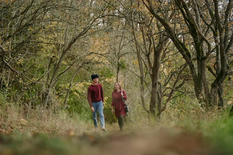 A candid portrait of a young couple walking along a quiet autumn woodland path, capturing genuine connection, warm tones and relaxed natural interaction with subtle authenticity.