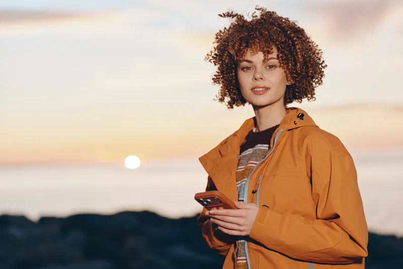 Woman with curly hair in orange jacket outdoors at sunset, holding phone and backpack strap, lifestyle travel portrait, warm coastal light, evening calm expression, beach horizon
