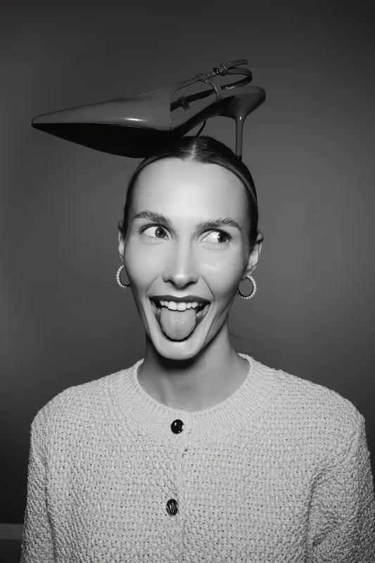 Happy white woman with light skin sticks out tongue playful with high heel shoe on head in studio.