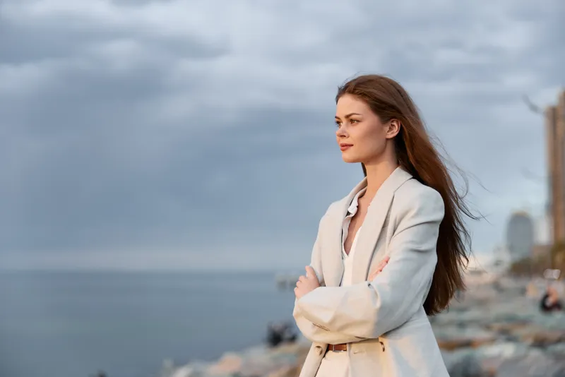 Confident businesswoman in a stylish blazer gazing at the horizon by the coastal cityscape, embodying ambition and strength against a dramatic sky.
