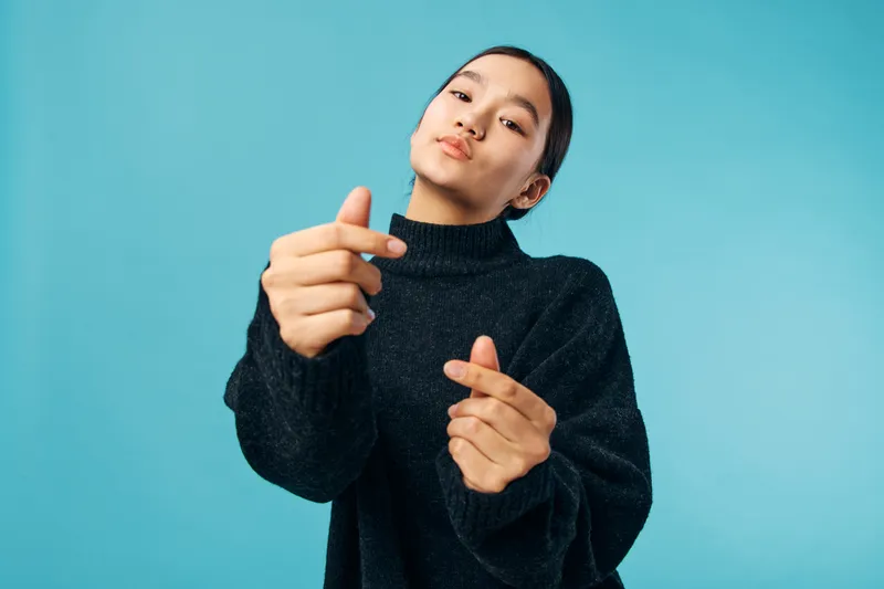 Young woman wearing black sweater making finger heart gesture with both hands, posing confidently against blue background in studio. Emotion and casual style with direct eye contact.