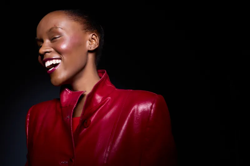 Smiling dark skinned African woman wearing red leather jacket looks away with closed eyes and bright lipstick on black background. Concept of joy, fashion, beauty, and confidence.