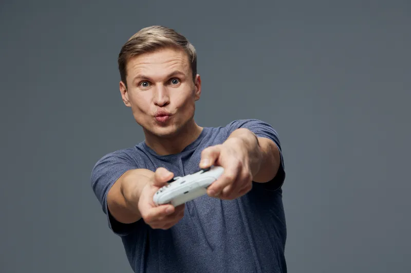 Smiling man playing with game controller, expressive facial expression, casual grey t-shirt, indoor setting, focused on gaming, youthful appearance, isolated on neutral background. People entertainmen