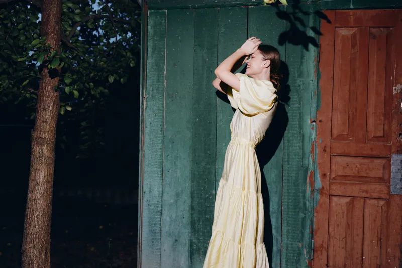 Woman in long yellow dress standing by wooden door in sunlight, summer portrait vintage mood, authenticity inclusivity